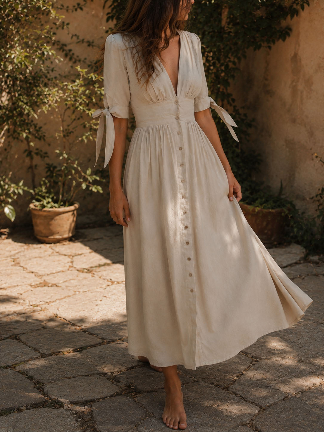 A woman wearing a cream-colored midi dress with a button-down front, tie details on the sleeves, and a V-neckline, standing barefoot on a stone patio in a sunny outdoor setting.
