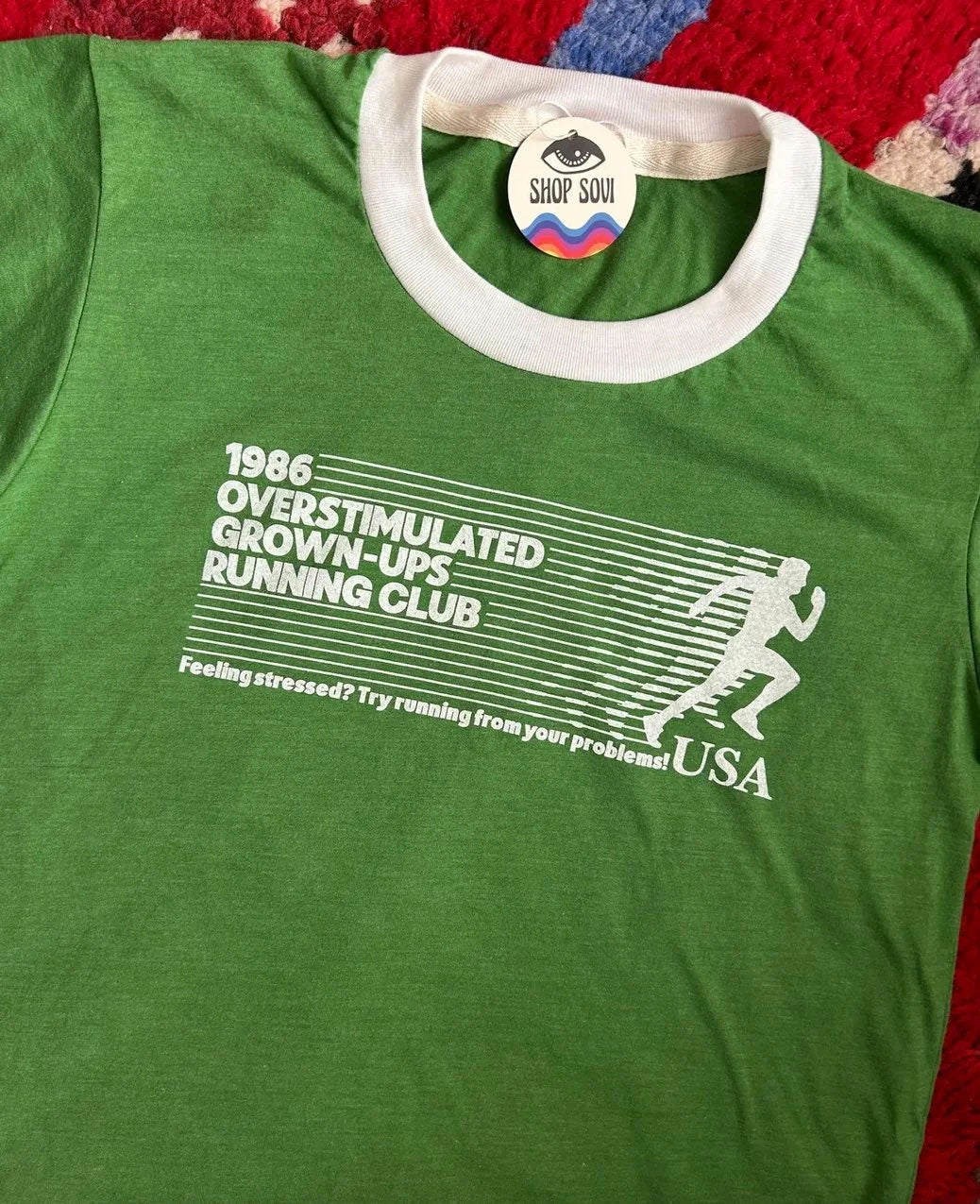 A green heather ringer t-shirt with a white ribbed crew neck and a white graphic print on the front reading '1986 OVERSTIMULATED GROWN-UPS RUNNING CLUB' with a running figure and 'USA'. A circular 'SHOP SOVI' brand tag is visible inside the collar.