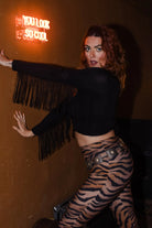 A woman posing in a dimly lit room wearing a black long-sleeve crop top with fringe and tiger-print leggings, next to a glowing orange neon sign that reads 'YOU LOOK SO COOL'.