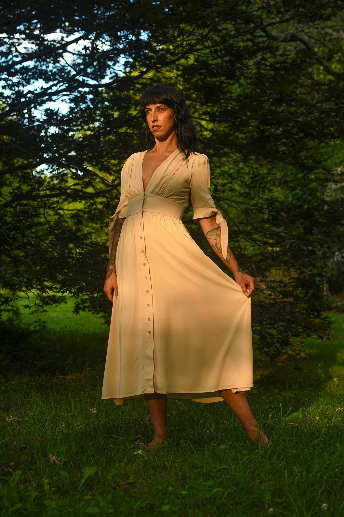 A woman wearing a cream-colored midi dress with a button-down front and three-quarter sleeves, standing barefoot in a grassy outdoor setting with trees in the background.