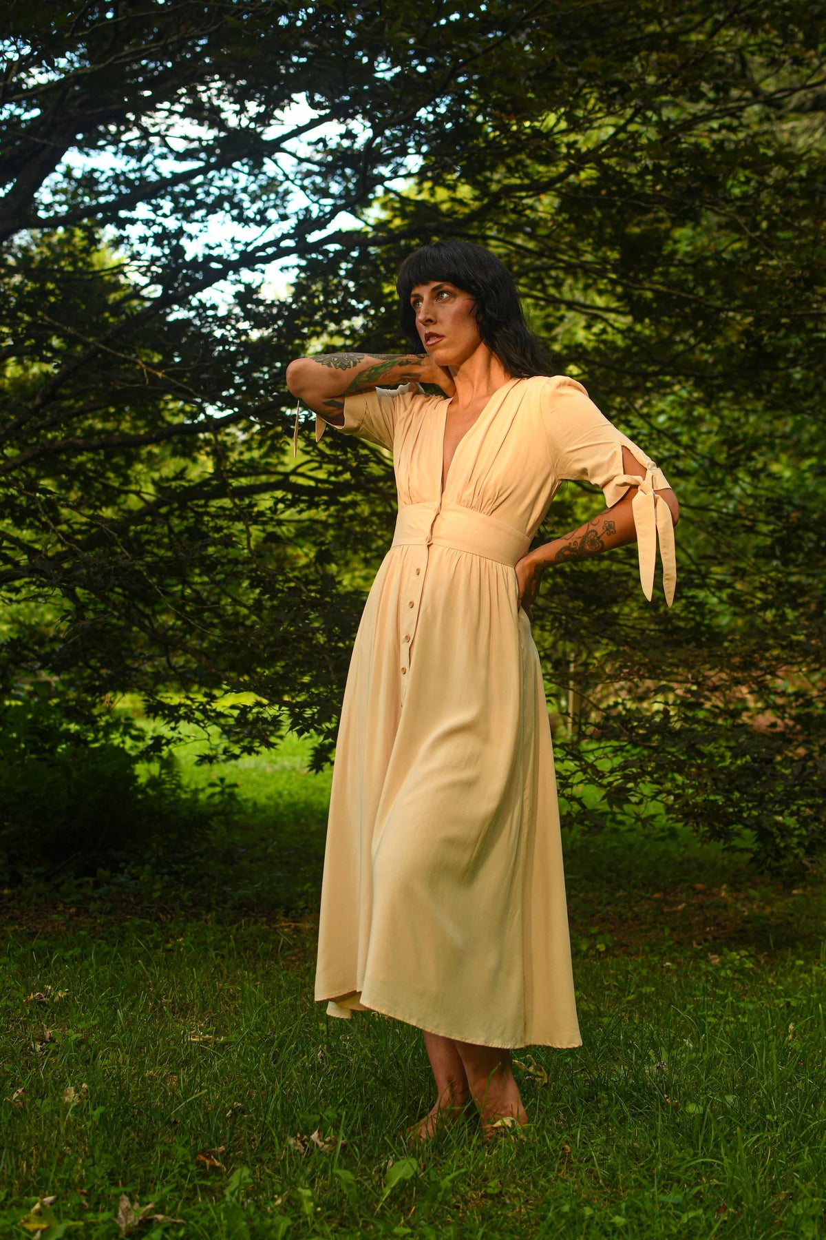 A woman wearing a light cream midi dress with tie details on the sleeves, standing outdoors in a grassy area with trees in the background.