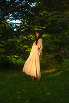A woman wearing a light beige maxi dress and a dark beanie stands on a grassy lawn surrounded by lush green trees in a park, backlit by warm golden sunlight.
