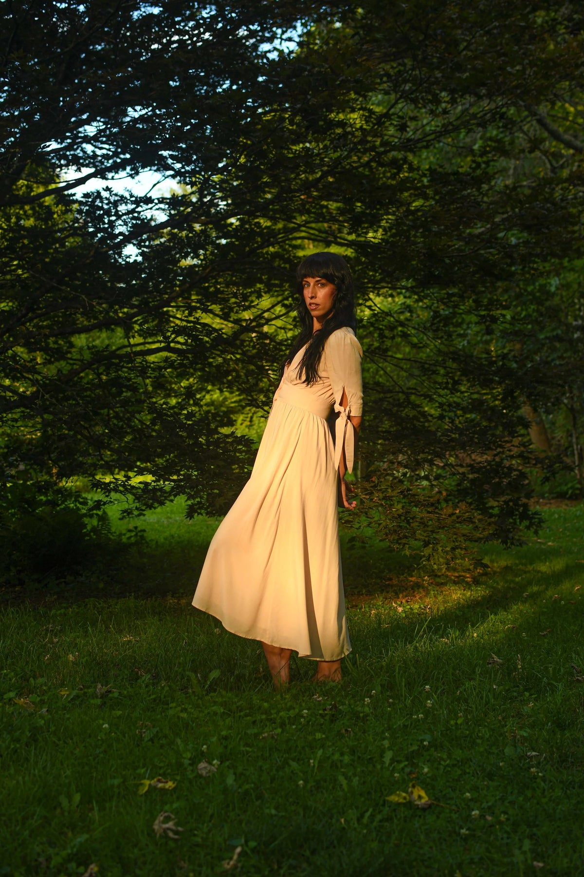A woman wearing a light beige maxi dress and a dark beanie stands on a grassy lawn surrounded by lush green trees in a park, backlit by warm golden sunlight.