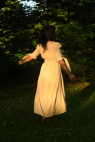 A woman wearing a cream-colored midi dress stands barefoot in a grassy forest clearing, viewed from behind as she looks up toward the trees.