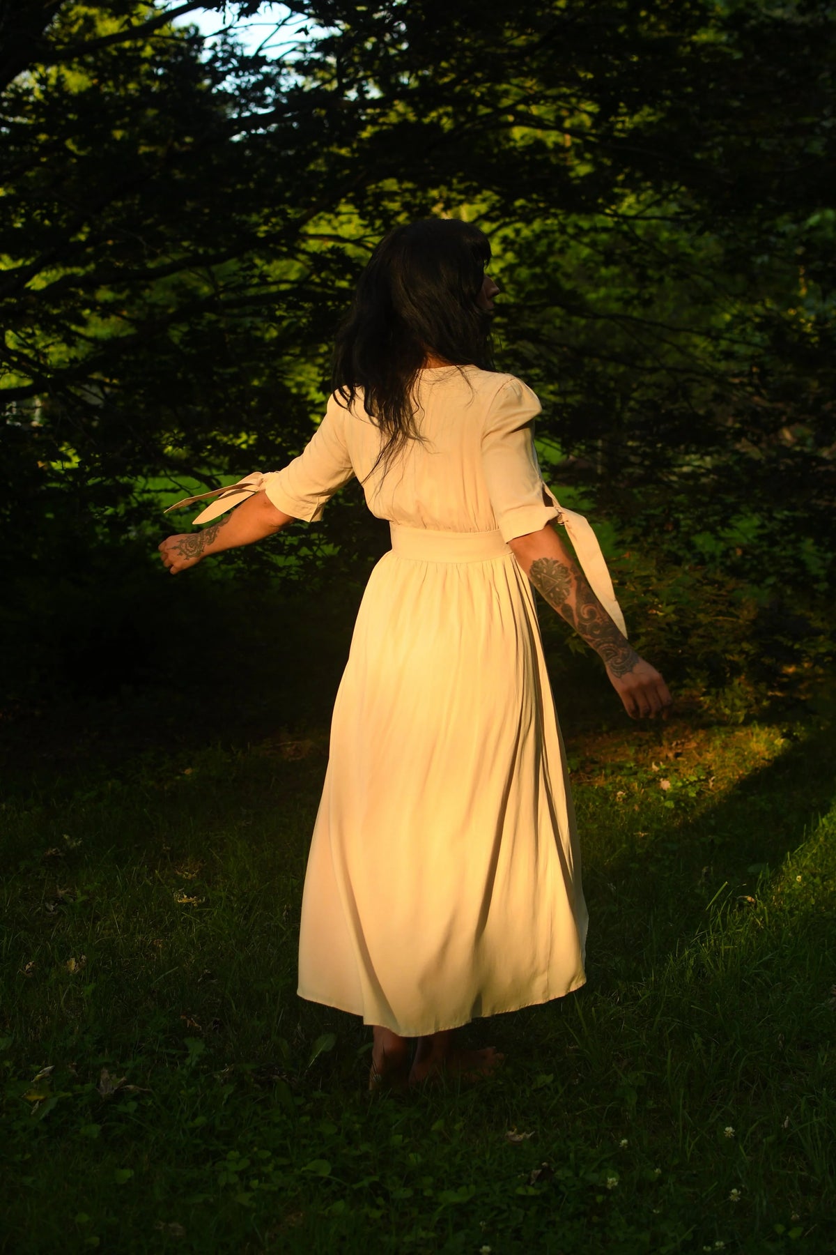 A woman wearing a cream-colored midi dress stands barefoot in a grassy forest clearing, viewed from behind as she looks up toward the trees.